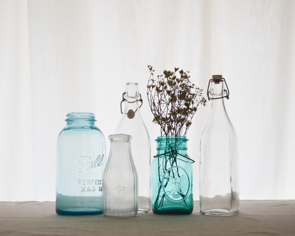 Des bouteille de plusieurs tailles, bleues et transparentes en contre-jour devant une fenêtre. L'une d'elles porte des fleurs séchées